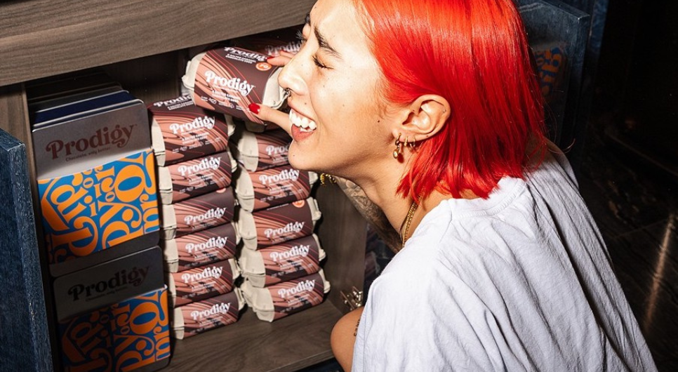 A woman with red hair reaches for an egg carton-style box of Prodigy's easter eggs from a stack of other boxes in a cabinet. She is laughing and wearing a white tea shirt.