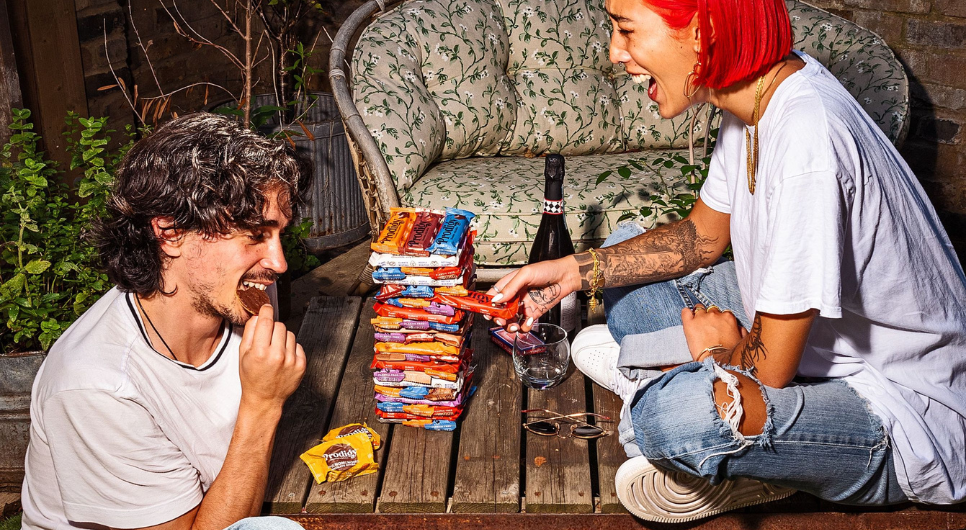 Two people - one with medium dark hair eating a chocolate biscuit and the other with bright red hair pulling a Prodigy chocolate bar from a Jenga-style stack - sit across from each other smiling.