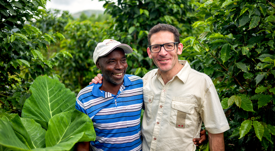 Two smiling men stand next to each other in a forest with an arm around the other