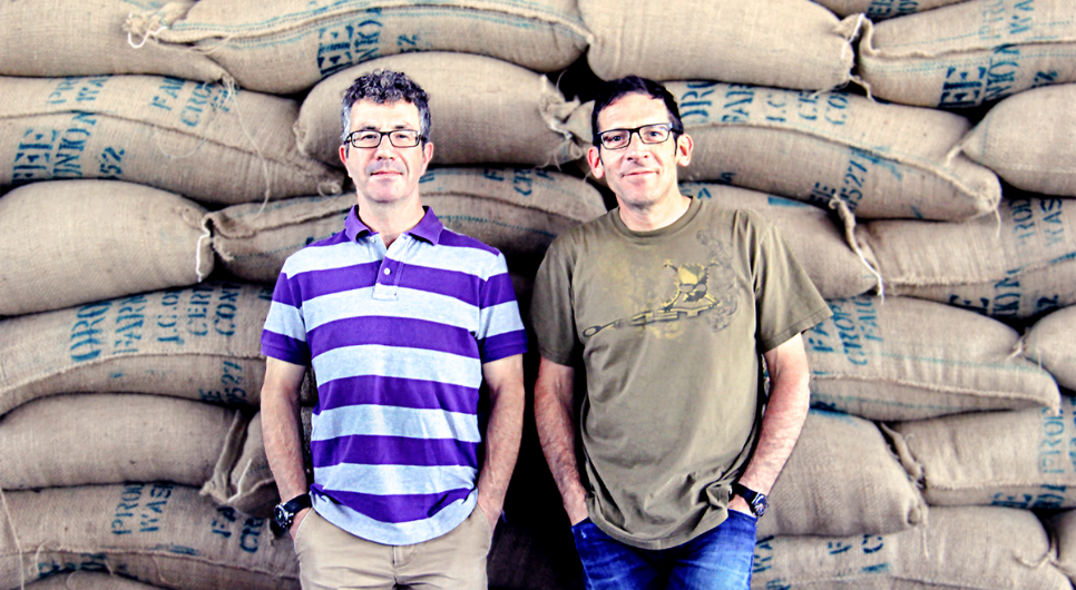 Two men wearing glasses stand next to each other in front of a tall pile of coffee bean sacks