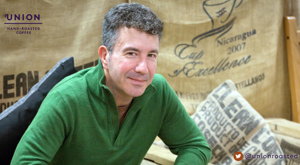 A man in a green shirt smiles at the camera, as he sits in front of sacks of yeast.