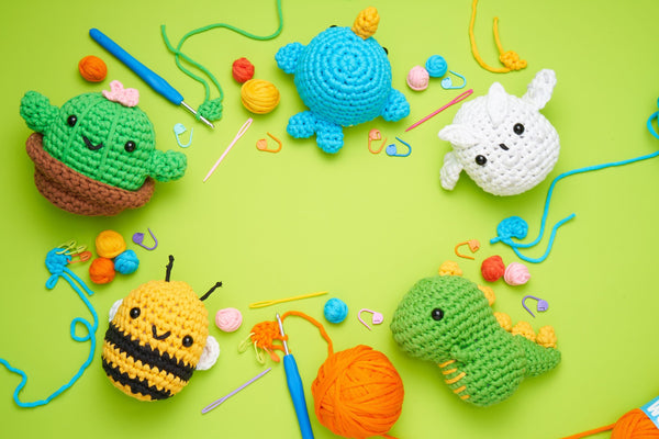 Beginner blue narwhal, white owl, green dino, yellow and black bee with white wings, and green and brown cactus on a green backdrop with crochet yarn, hook and stitch markers between them