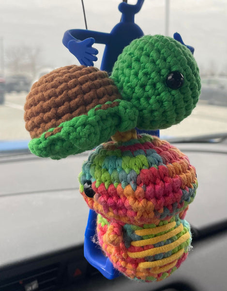 A turtle and dinosaur crochet plushie hanging on a car's front mirror