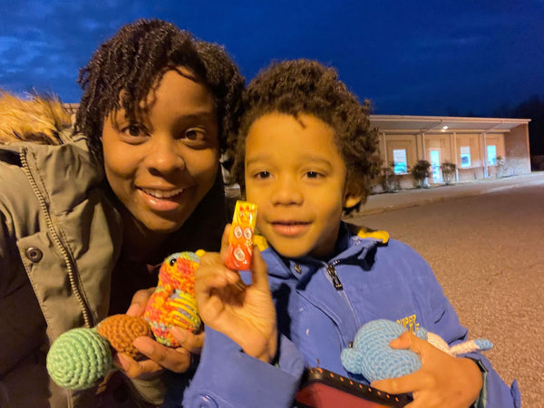 An African-American woman holding dinosaur and turtle crochet plushies, standing next to her son who's holding penguin and yeti plushies