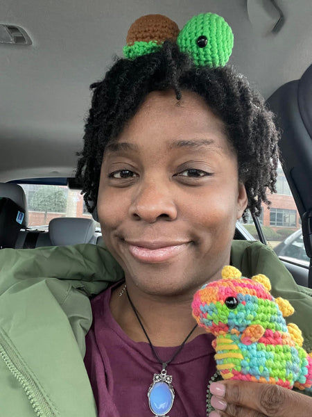 An African-American woman in a car holding a multi-colored dinosaur crochet plushie in her right hand and a turtle crochet plushie on her head