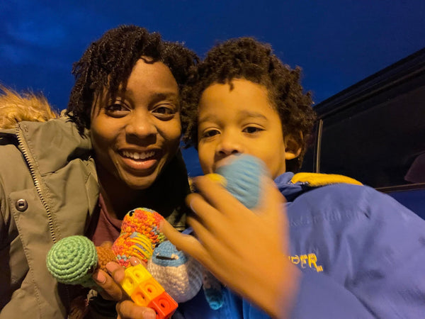 An African-American woman holding dinosaur and turtle crochet plushies, standing next to her son who's holding penguin and yeti plushies