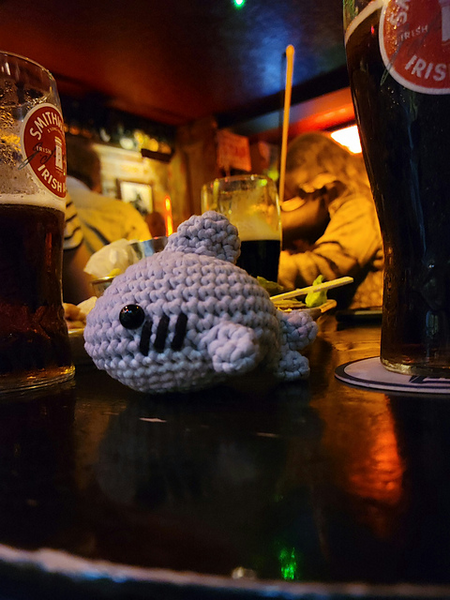 A shark crochet plushie on a table in between two beer glasses