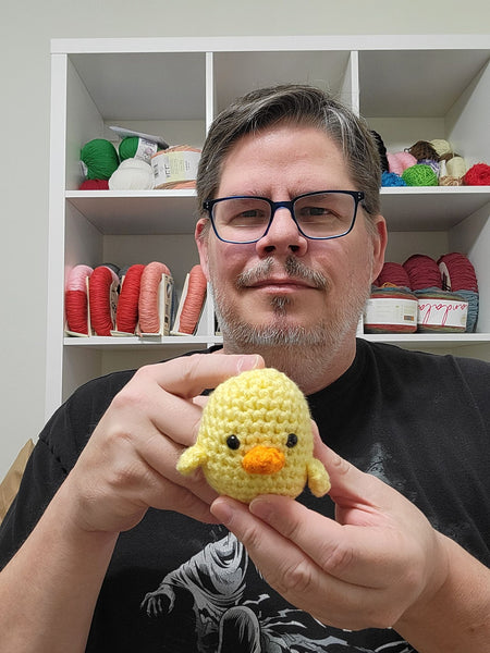 A white man in-front of a cabinet with yarn wearing a black T-shirt and holding a yellow chick amigurumi plushie