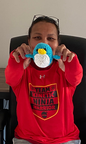 A woman in a red T-shirt holding out a blue penguin crochet plushie