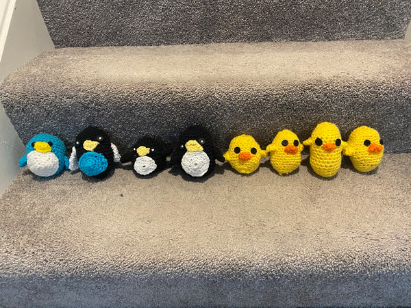 Four penguin crochet plushies, one blue and three black, and four yellow chick crochet plushies lined up on a brown, carpeted stair step