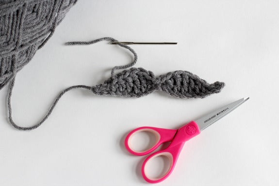 A gray moustache with a tapestry needle and yarn attached to it, and a scissor with pink handles next to it