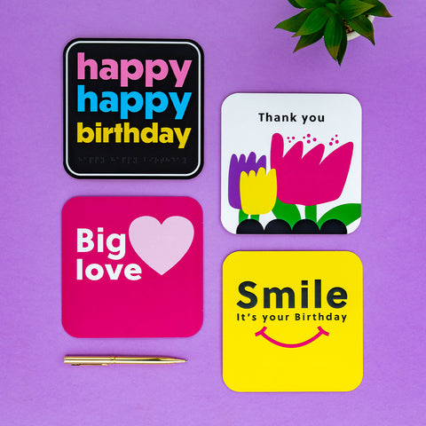 A purple background with a plant peaking through the top right, and a golden pen horizontally lay on the bottom left, there are 4 cards layout, a black happy birthday card, a white thank you card with flowers, a pink 'big love' card with a heart and a yellow birthday card with a smile and birthday message.