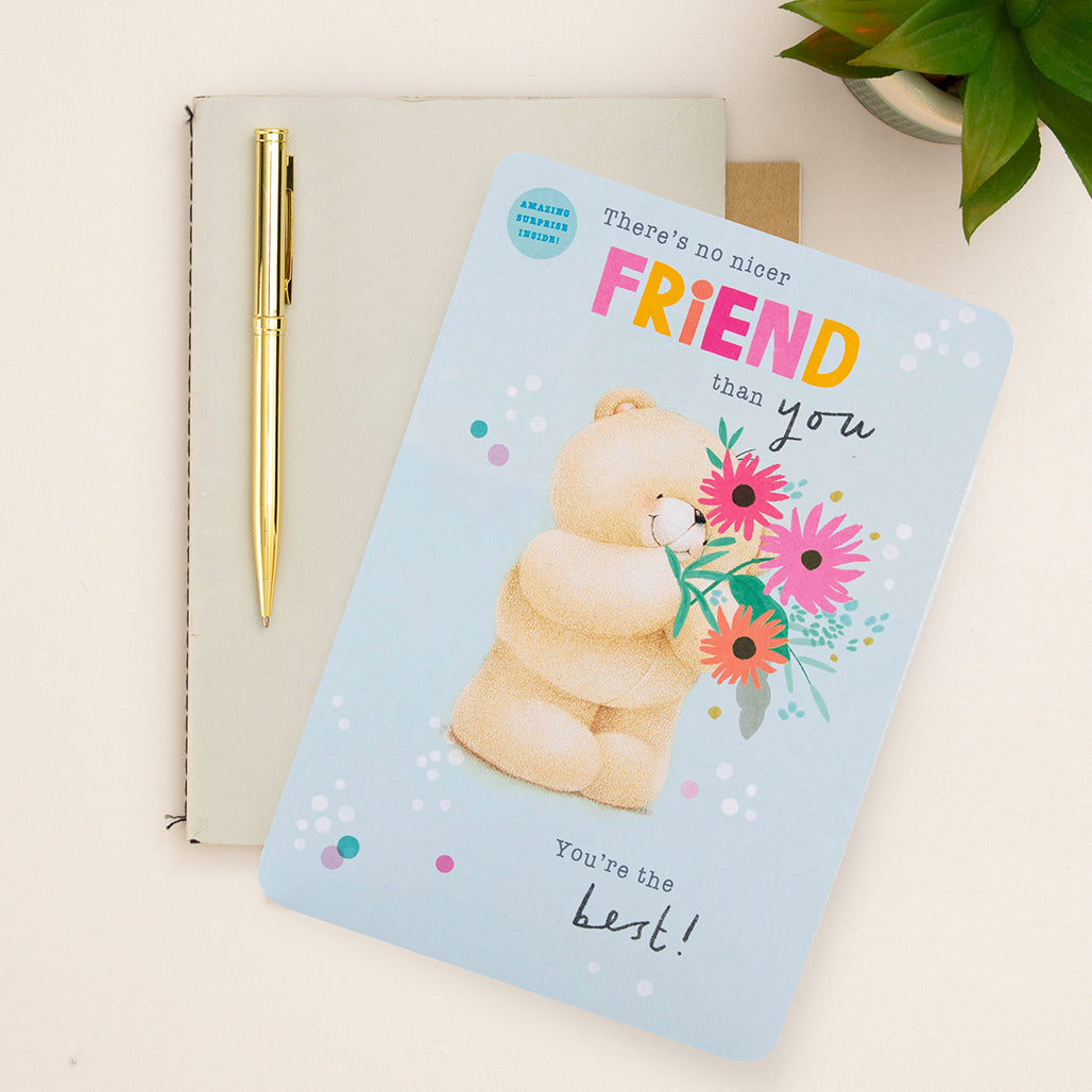 A neutral background, half a green plant in the corner, with a beige notebook, a gold pen, and a blue Forever Friends card for a friend, with a bear holding flowers. 