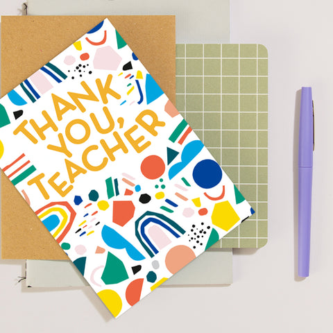 A neutral coloured square background, with a slightly darker flat notebook, a green square patterned paper, and a cork textured notebook on top, with a funky patterned colourful Thank you teacher card on top on an angle and a purple pen on the right lying straight.