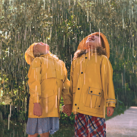 Two girls stood outside holding hands, both girls are in a yellow rain coat, their faces turned towards the sky and both are trying to catch raindrops in their mouth, the background is a green hedge with rain falling heavily all around.