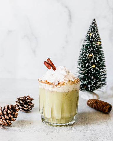 Photo of egg nog made with matcha green tea powder, with festive Christmas tree and pine cone decorations in background.