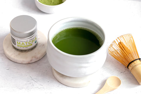 Photo of a small silver tin of Sencha Naturals Ceremonial Grade Matcha Powder on the left, a small white cup of liquid green tea in the center, and a small wooden spoon and bamboo whisk on the right, all on a white background