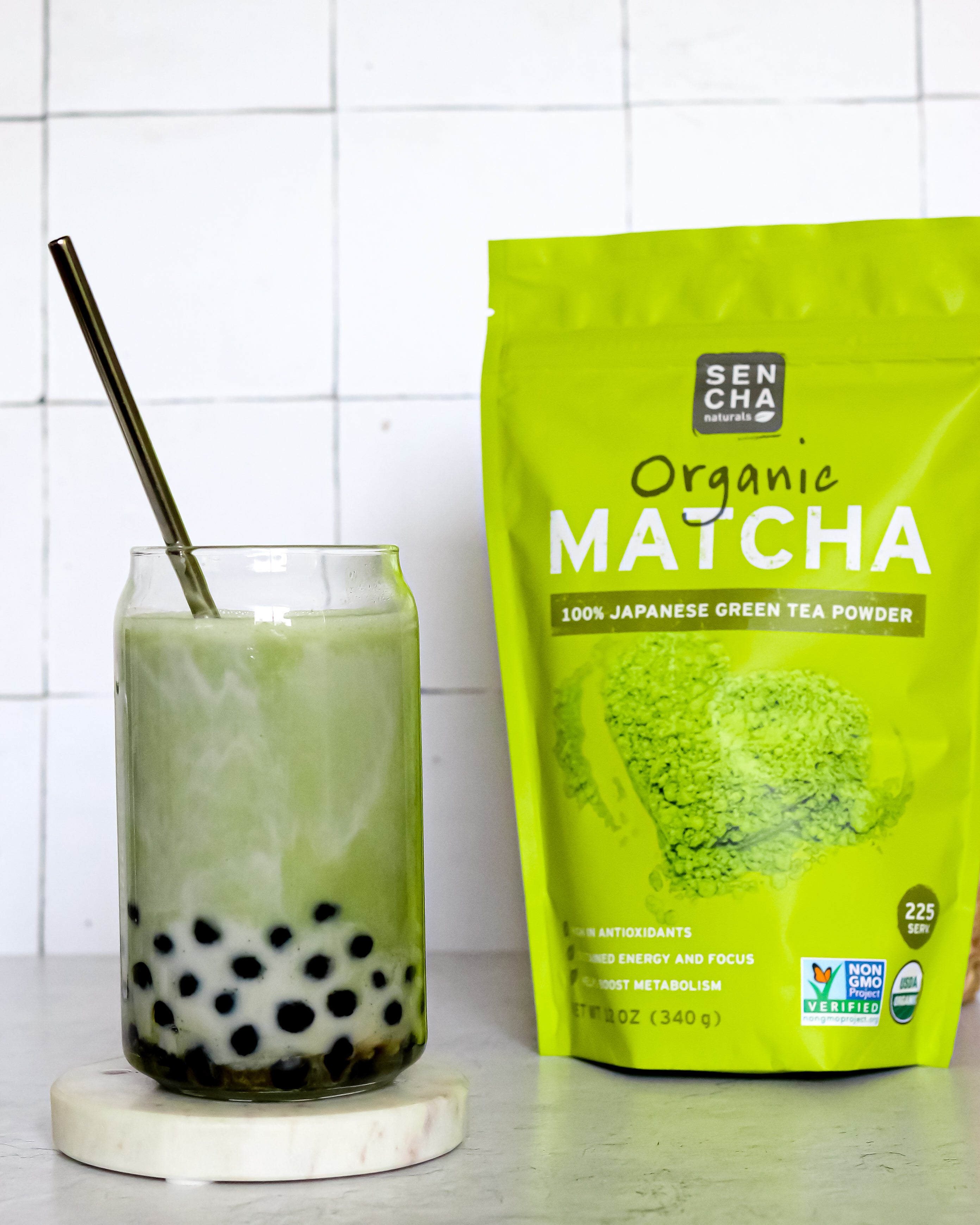 Photo of a bag of sencha naturals matcha green tea powder on the right, a clear glass of green tea boba bubble tea on the left with a silver straw inside, all on a white background