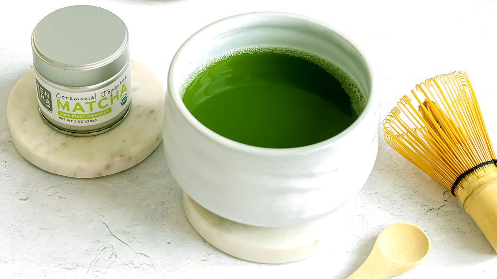 Photo of a small silver tin of Sencha Naturals Ceremonial Grade Matcha Powder on the left, a small white cup of liquid green tea in the center, and a small wooden spoon and bamboo whisk on the right, all on a white background