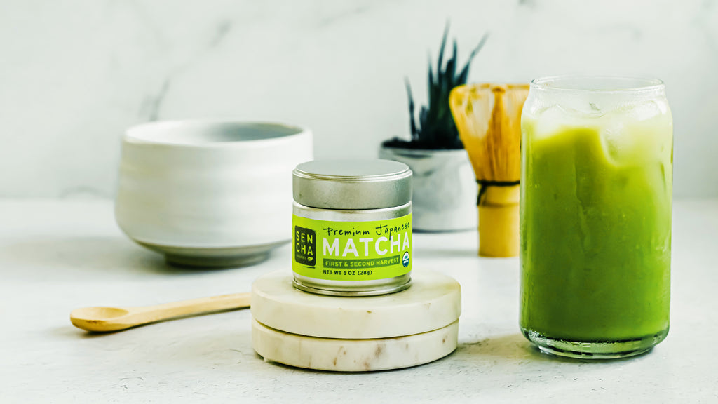 Photo of Sencha Naturals Premium Matcha tin, a small white cup, and an Iced Latte in a clear glass, all on a white background with a bamboo whisk and a green plant in the back