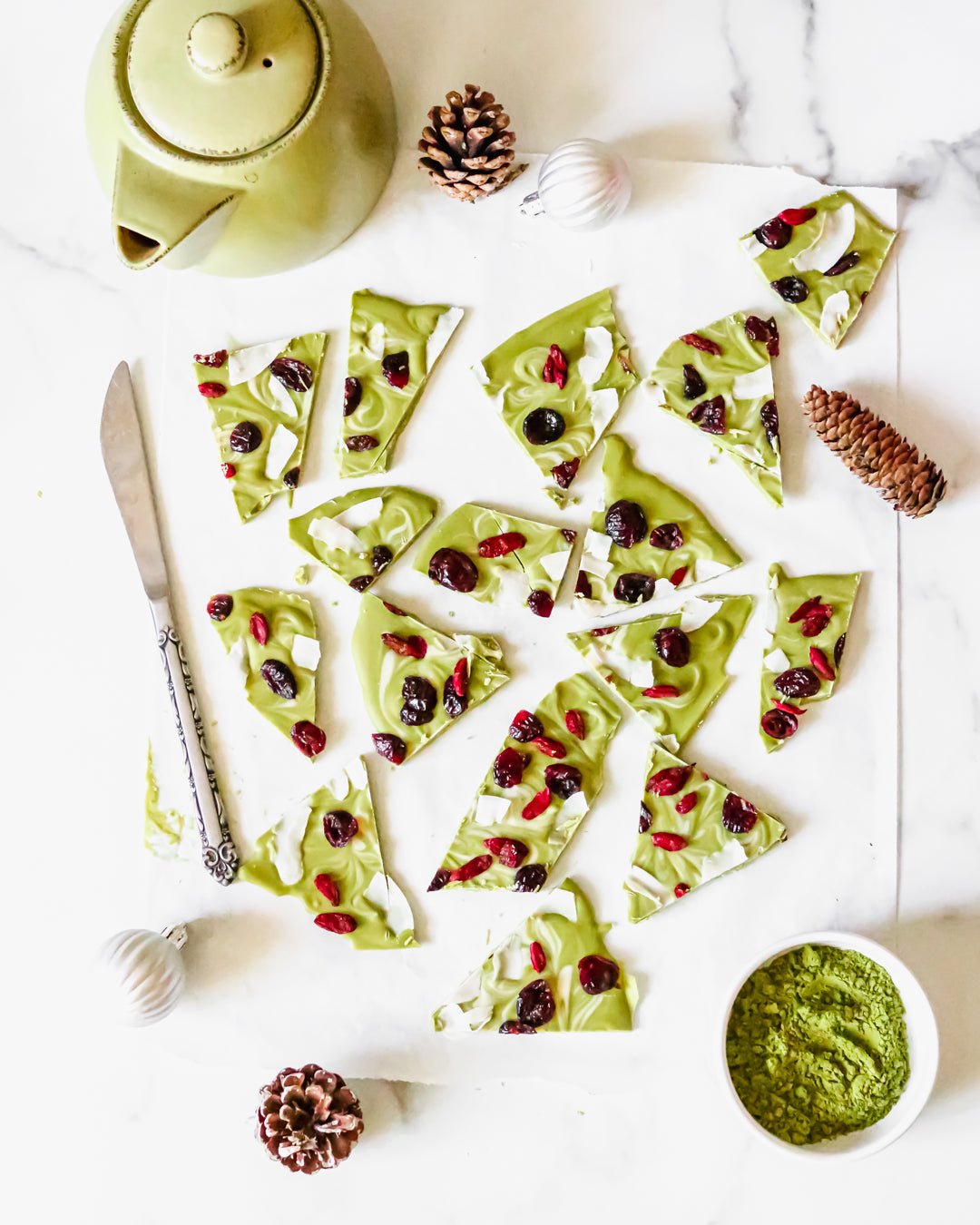 Photo of several pieces of green tea Holiday Bark dessert treats, topped with cranberries and white coconut strips, surrounded by a few decorative pine cones, a small green teapot, a small bowl containing matcha green tea powder, a silver butter knife, and two white Christmas globes, all on a white background