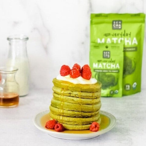 Photo of a stack of green tea pancakes covered with raspberries and maple syrup on a white plate, with a bag of Sencha naturals matcha green tea powder in the back, a clear jar of milk in the back left, a small clear bottle of maple syrup next to the milk, all on a white background