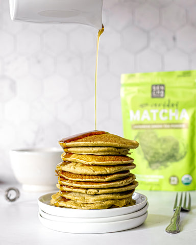 Photo of a stack of pancakes made with matcha green tea powder, with maple syrup being poured on top, and a bag of matcha green tea in the background, all set on a white countertop.