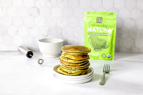 Photo of a stack of pancakes made with matcha green tea powder, with a silver fork on the right, a small silver electric whisk and white bowl in the back, and a bag of matcha green tea in the background, all set on a white countertop.