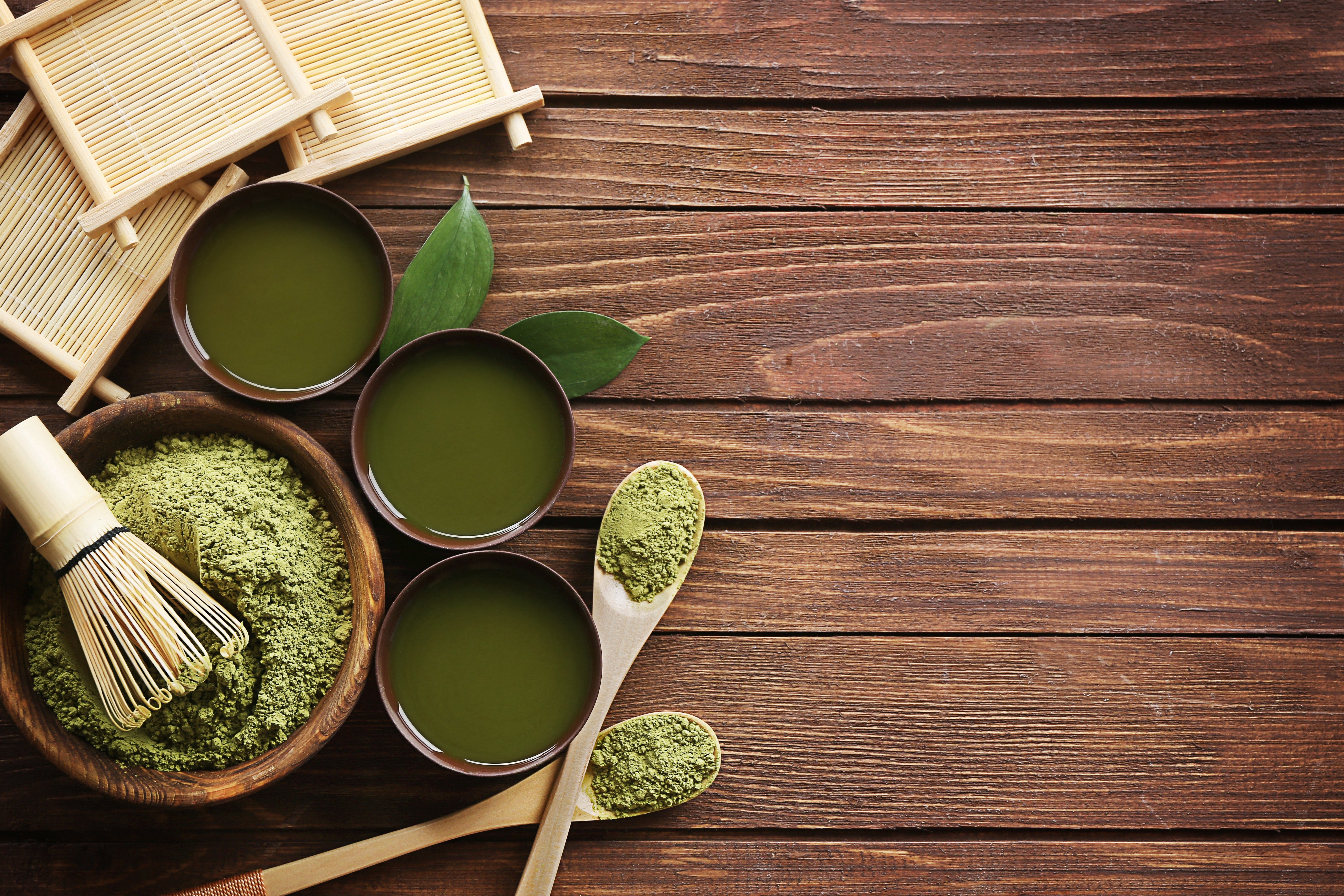 Photo of a small bowl containing green tea powder with a bamboo whisk on top, 2 small wooden spoons to the right, 3 light wooden coasters, and 3 cups of liquid green tea all on a dark wooden tabletop
