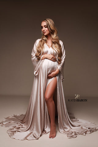 Blondes schwangeres Model posiert in einem Studio und trägt ein langes, seidiges Kleid in Sandfarbe. Das Kleid hat einen Schlitz an der Seite, wo eines der Beine des Models zu sehen ist. Sie hält ihren Bauch mit beiden Händen.