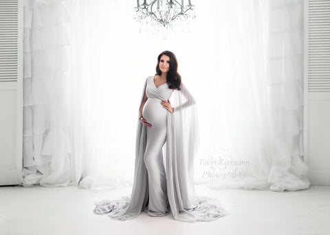 pregnant model with long dark hair poses in a studio wearing a dress in cool grey color. the dress is made of jersey and chiffon. the dress features a short sleeves neckline with an adjustable sweetheart top. long chiffon sleeves are attached to the short sleeves and can be worn as a cape. the dress is long enough to cover the feet from the model. 