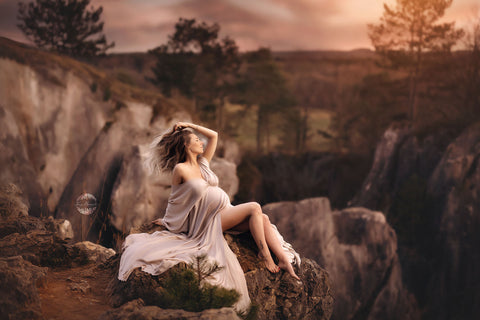 pregnant model sitting on rocks outdoor wearing a sand long maternity dress made of silk. 