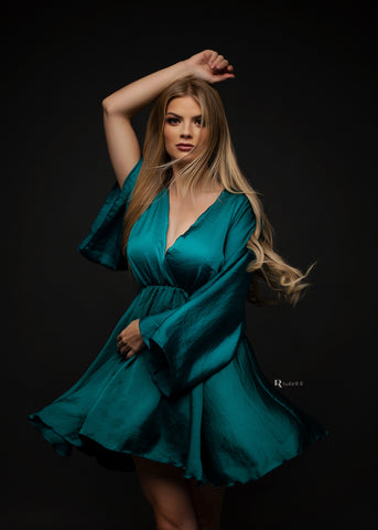 blond model poses in a dark studio wearing a petrol color silky dress. the dress is short and has a low v cut neckline. 