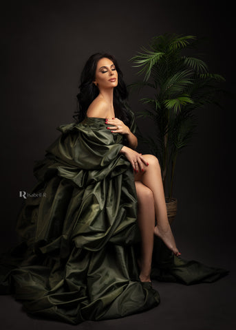 Brunette model poses sitting on chair against a dark brown background in a studio next to a green plant. she is wearing a taffeta dress in dark green color. 