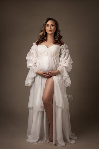 brunette model poses in a studio in front of a brown background. she is wearing a chiffon dress in off white. 