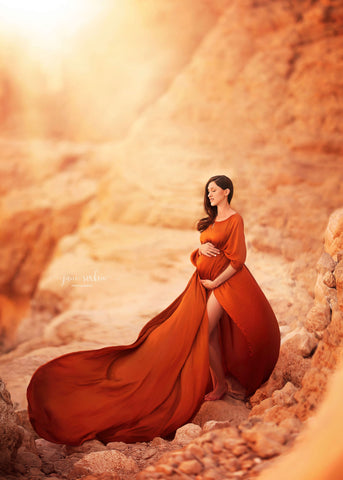 brunette model poses outdoors during a maternity photoshoot wearing a long cape in cognac color. the cape is made of silk and has on shoulder neckline. she has her eyes closed and holds her belly while posing. she is standing by a light brown rocky/ sandy place. 