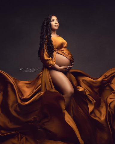 Séance photo de maternité en studio d'un modèle aux cheveux noirs portant une tenue cape soyeuse de couleur cognac. elle pose avec la cape un peu ouverte de manière à montrer partiellement sa jambe et son ventre.