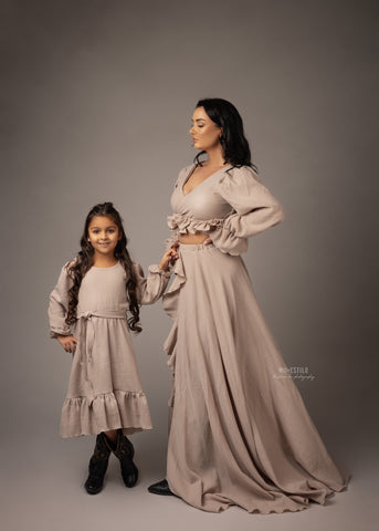 Mutter und Tochter posieren Händchen haltend in einem Studio. Beide tragen ein sandfarbenes Kleid im Boho-Stil.