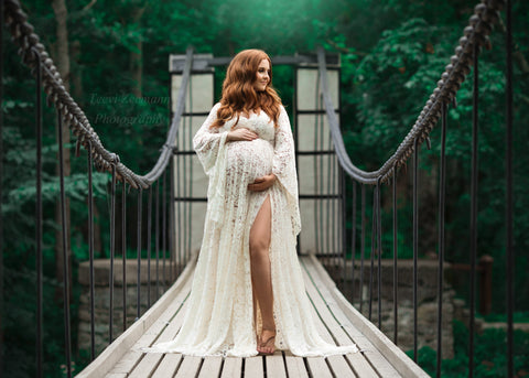 Modèle enceinte aux cheveux rouges pose au milieu d'un pont vêtue d'une robe de maternité blanc cassé entièrement en dentelle.