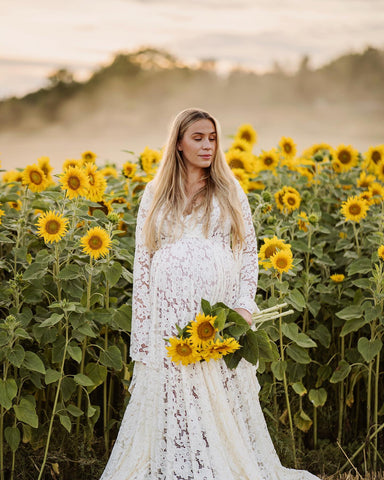 Blondes schwangeres Model posiert neben einem Sonnenblumenfeld und trägt ein weißes Spitzenkleid