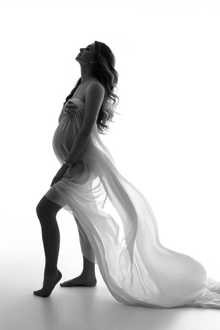 Schwarz-Weiß-Foto eines schwangeren Models, das in einem Studio vor weißem Hintergrund posiert. Sie trägt einen Chiffonschal in gebrochenem Weiß, der ihren Körper teilweise bedeckt.
