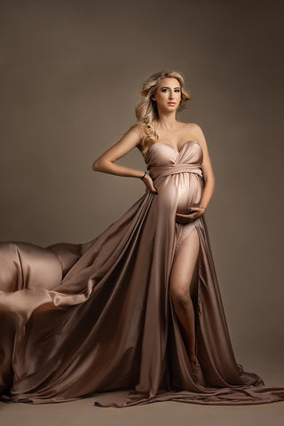 Modèle enceinte blonde pose dans un studio vêtue d'une longue robe soyeuse avec une fente sur le côté de la jupe.