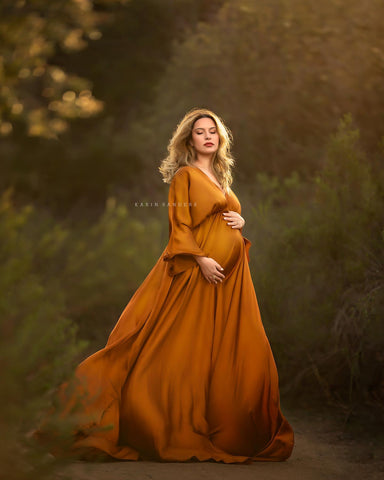 Blondes schwangeres Model posiert während der goldenen Stunde draußen vor vielen Bäumen. Sie trägt ein langes, seidiges Kleid in Cognacfarbe.