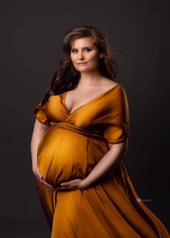 Brünettes schwangeres Plus-Size-Model posiert in einem Studio mit dunklem Hintergrund und trägt ein seidiges cognacfarbenes Kleid. Das Kleid hat einen tiefen V-Ausschnitt und bedeckt einen Teil ihres Arms. Das Model hält ihren Bauch mit beiden Händen.