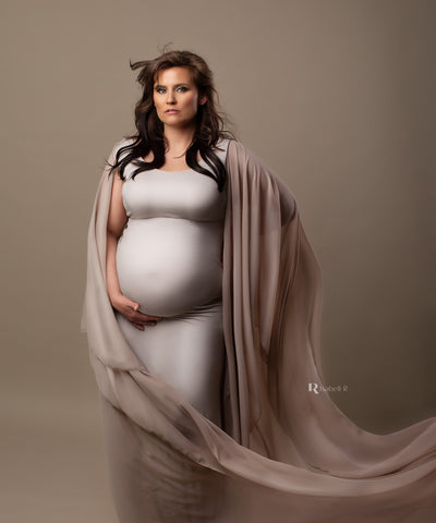 plus size brunette model poses in a studio wearing a long plus size maternity dress in sand color. she holds her bump with one hand and the other hand and arm are covered by the flattering chiffon sleeves from the gown. the sleeves are long and sewed as a cape. the model looks straight to the camera and her dress is in sand color.