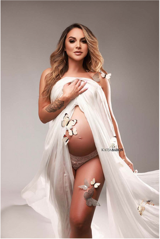 Dunkelblondes schwangeres Model posiert in einem Studio, trägt einen seidenen Schal auf der Brust und lässt Bauch und Beine unbedeckt. Dem Foto wurden digitale weiße Schmetterlinge hinzugefügt.