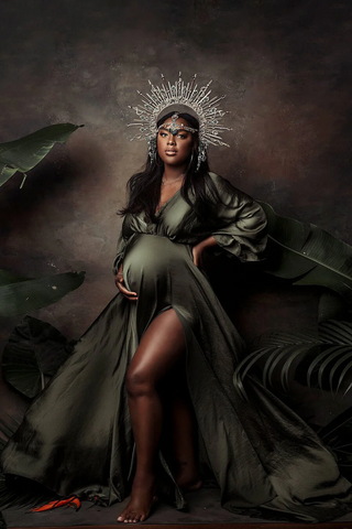 black pregnant model poses in a studio next to a few green plants wearing a stunning long dress made of silk in olive color. she has a crown to match the style. 