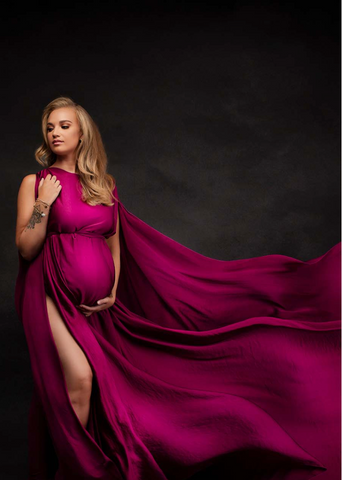 Blondes schwangeres Model posiert während eines Schwangerschaftsfotoshootings mit geschlossenen Augen in einem Studio. Sie trägt einen kirschroten seidenen Umhang mit schulterfreiem Oberteil und viel herunterfallendem Stoff.