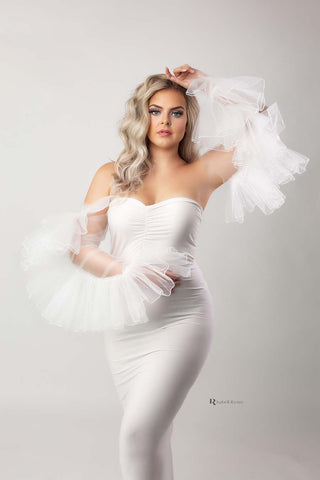 blond model poses in a white studio wearing a white outfit. she wears a tight white strapless dress and has tulle ruffled sleeves to match the style.