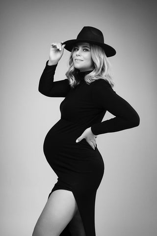 Schwangeres Model posiert in einem Studio und trägt ein schwarzes langes Kleid mit langen Ärmeln und Rollkragen. passend zum Stil trägt sie auch einen schwarzen Hut.
